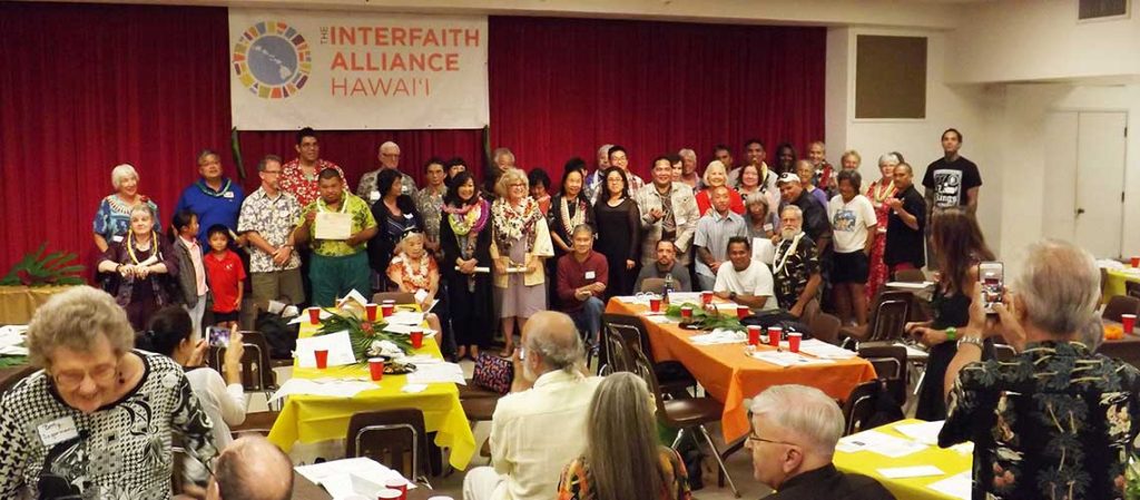 many smiling people grouped near the stage under The Interfaith Alliance Hawaii banner